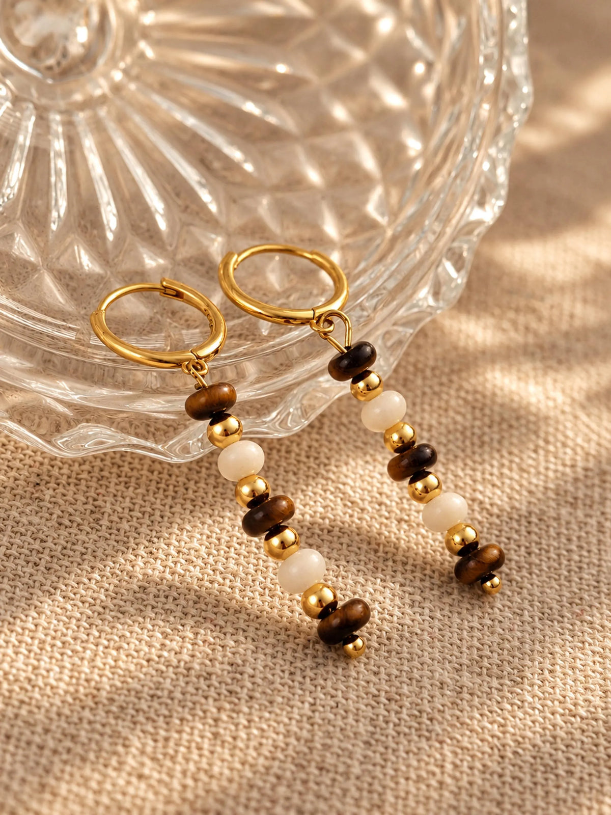 Tiger's Eye and Mother of Pearl beaded gold drop earrings displayed on a ribbed glass bowl and linen surface.