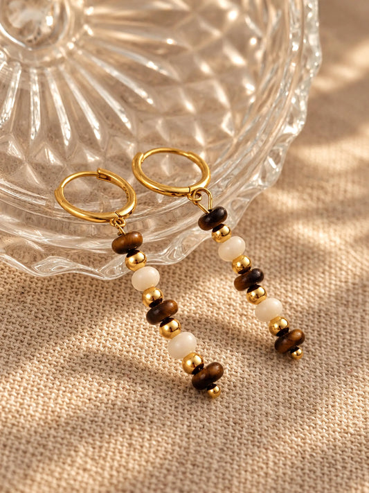 Tiger's Eye and Mother of Pearl beaded gold drop earrings displayed on a ribbed glass bowl and linen surface.