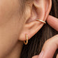 A close-up of a woman's ear wearing a gold hoop earring and a thin white enamel gold ear cuff on cartilage.