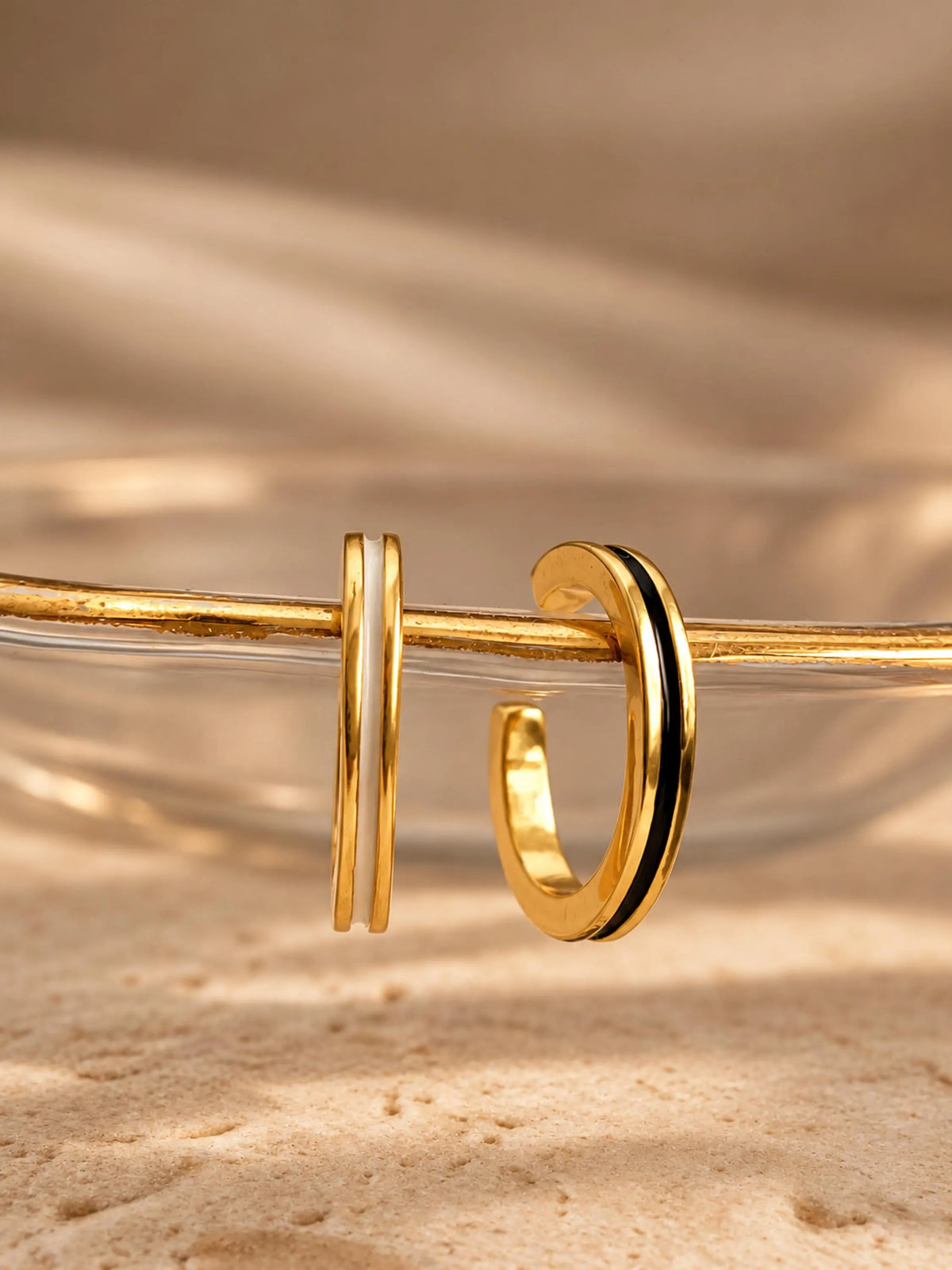 2 gold ear cuff earrings displayed on a clear glass tray over a sandy surface, one in black enamel and one in white enamel with warm beige background lighting.