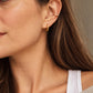 A close-up of a woman's ear wearing a small gold hoop earring and a thin gold ear cuff, with her brown hair and white tank top visible against a soft neutral background.