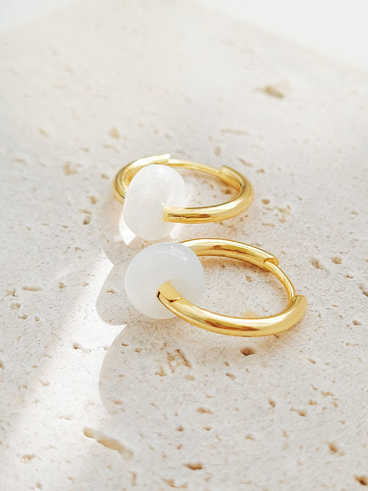 A pair of luxurious white jade huggie hoop earrings crafted in yellow gold vermeil displayed atop a sleek marble surface, highlighting their genuine gemstone allure.