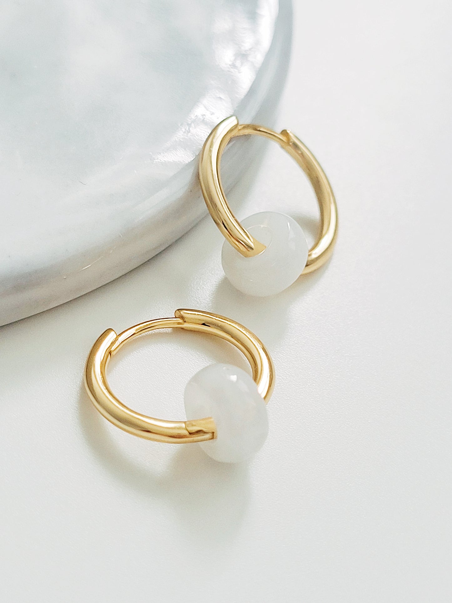 Two stunning white jade huggie hoop earrings gracefully positioned on a marble surface, emphasizing their elegant craftsmanship. Earrings are crafted in 18ct gold plated on silver.