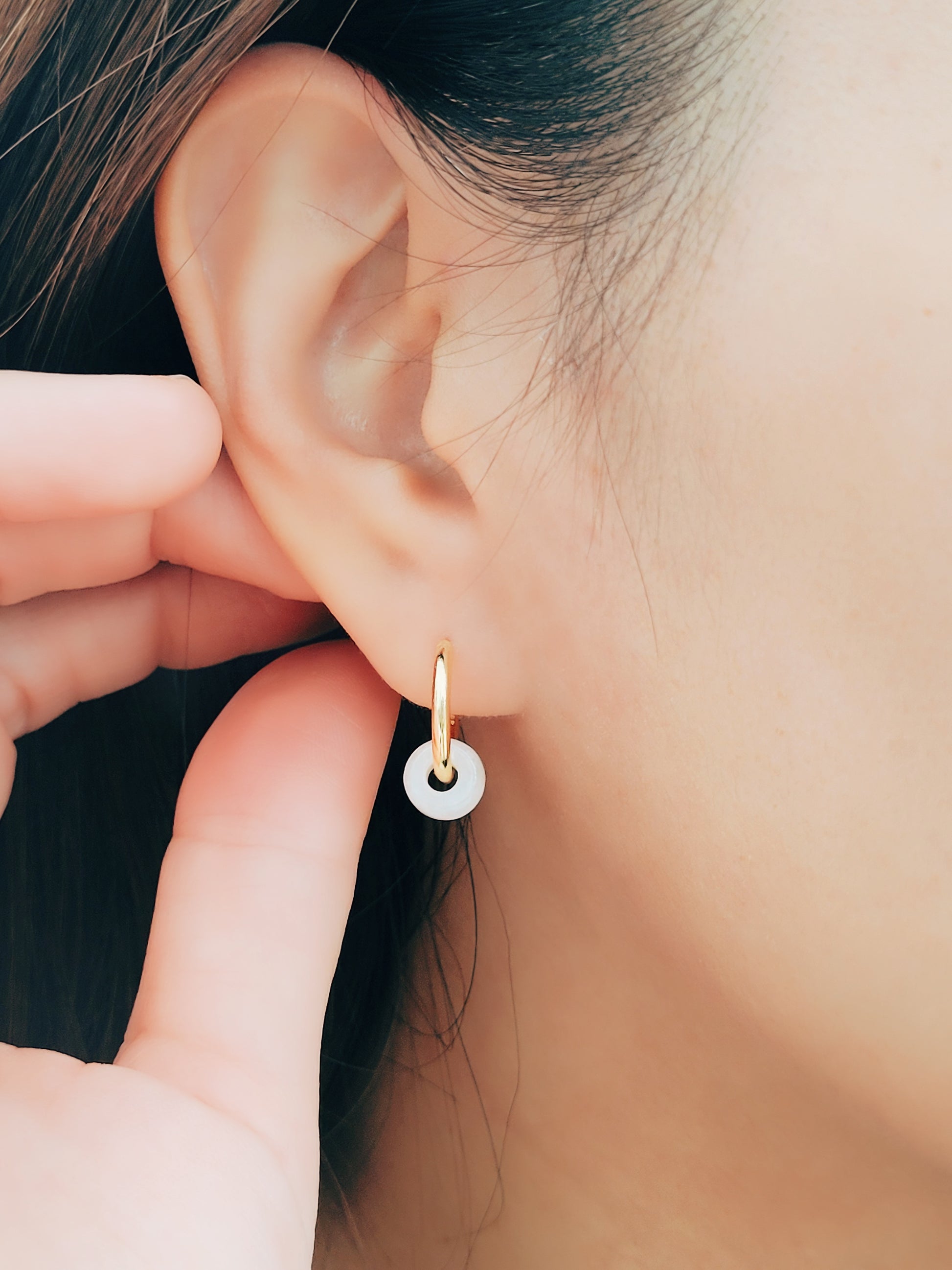 A woman adorns her ear with a small gold jade earring, complemented by white jade huggie hoops made from natural gemstones.