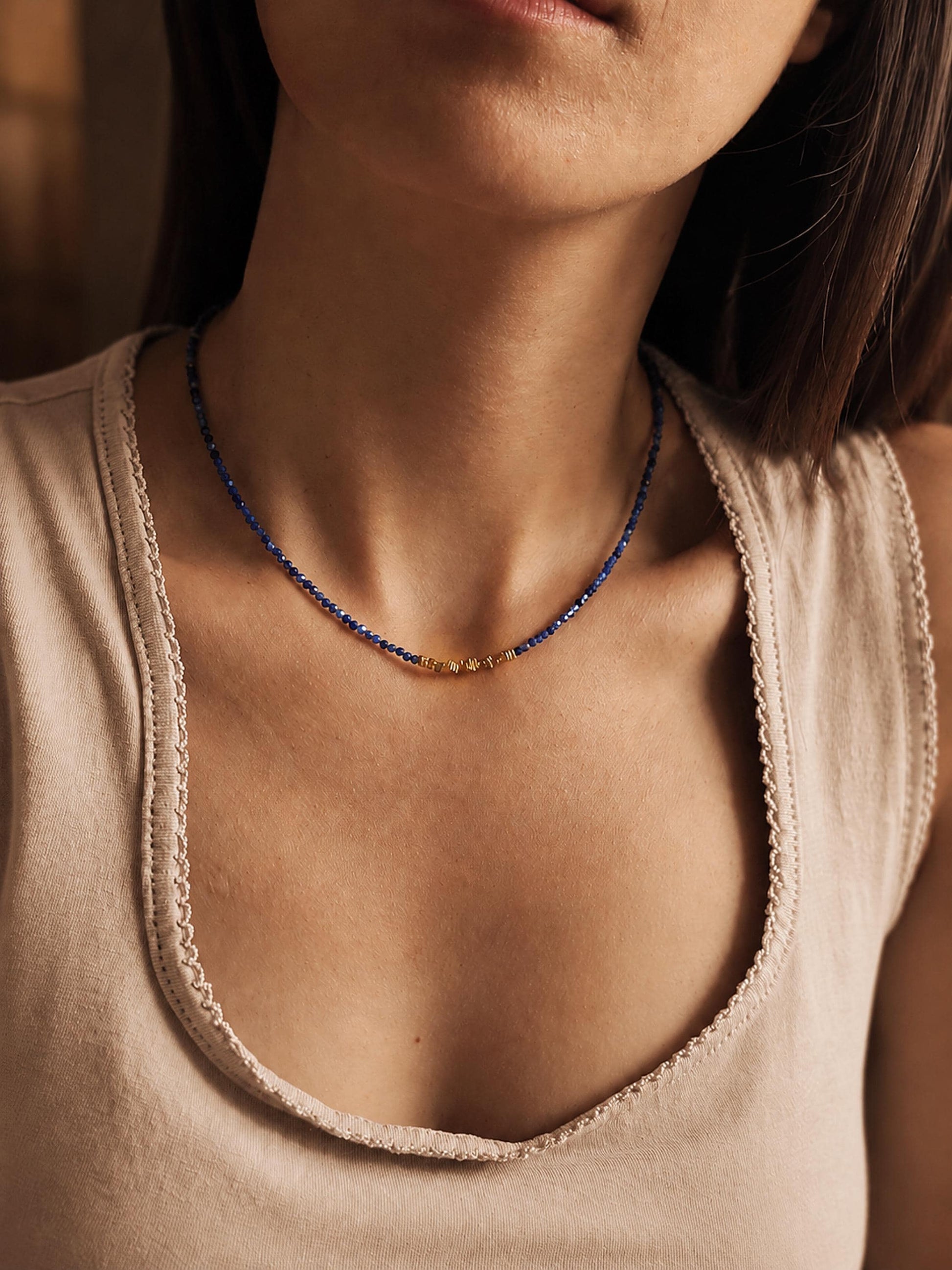 A close-up lifestyle shot of a model wearing a delicate Kyanite beaded necklace. The necklace features small blue beads with a central gold accent and a small gold pendant.