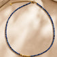 A delicate beaded necklace with deep blue faceted kyanite gemstone beads and a textured 22k gold vermeil bead at the center, displayed on a beige satin fabric background.
