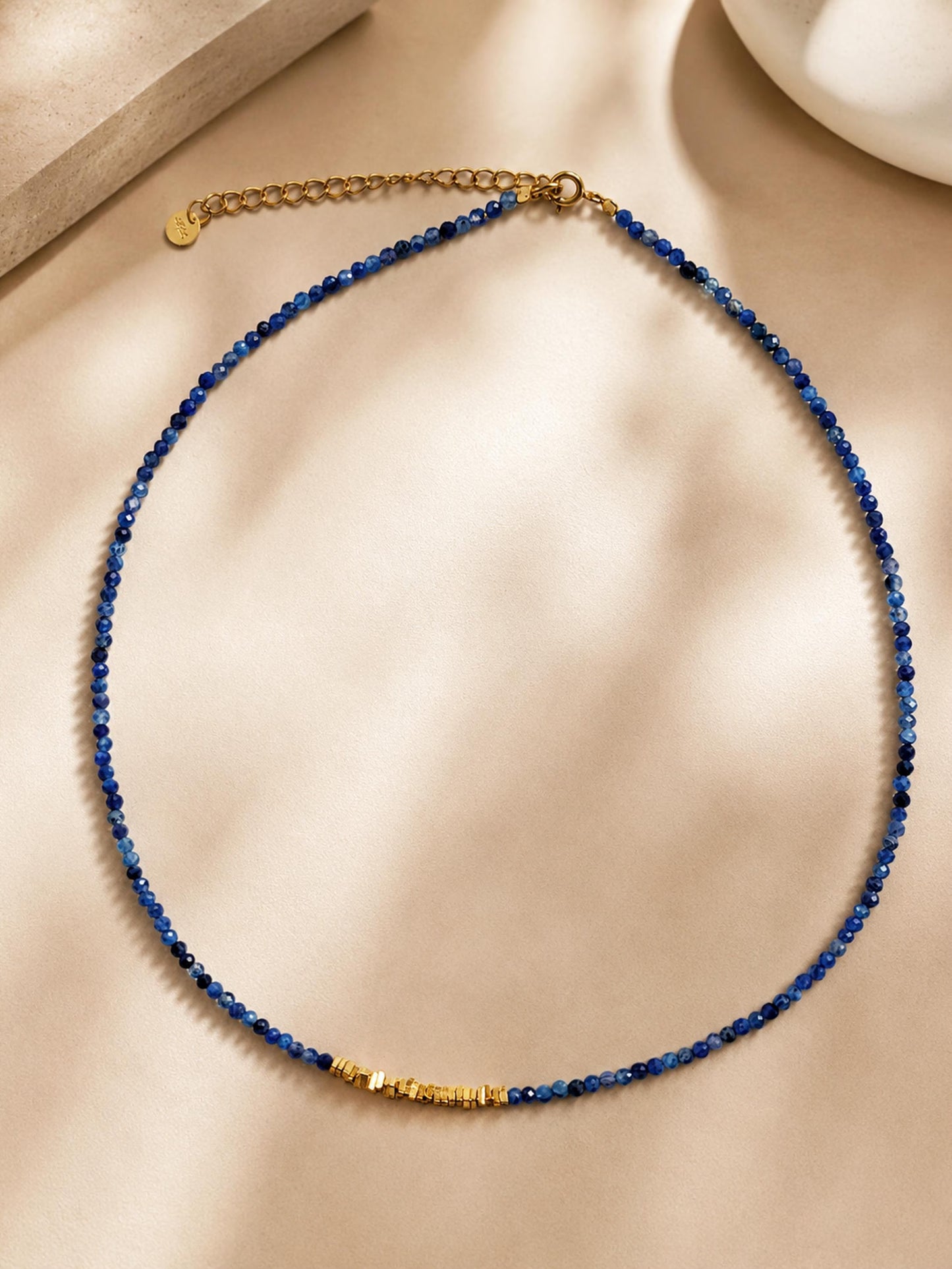 A delicate beaded necklace with deep blue faceted kyanite gemstone beads and a textured 22k gold vermeil bead at the center, displayed on a beige satin fabric background.