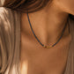 A close-up lifestyle shot of a woman wearing a delicate royal blue beaded necklace with dark blue kyanite beads and a central gold-toned bead accent, paired with a beige satin top.