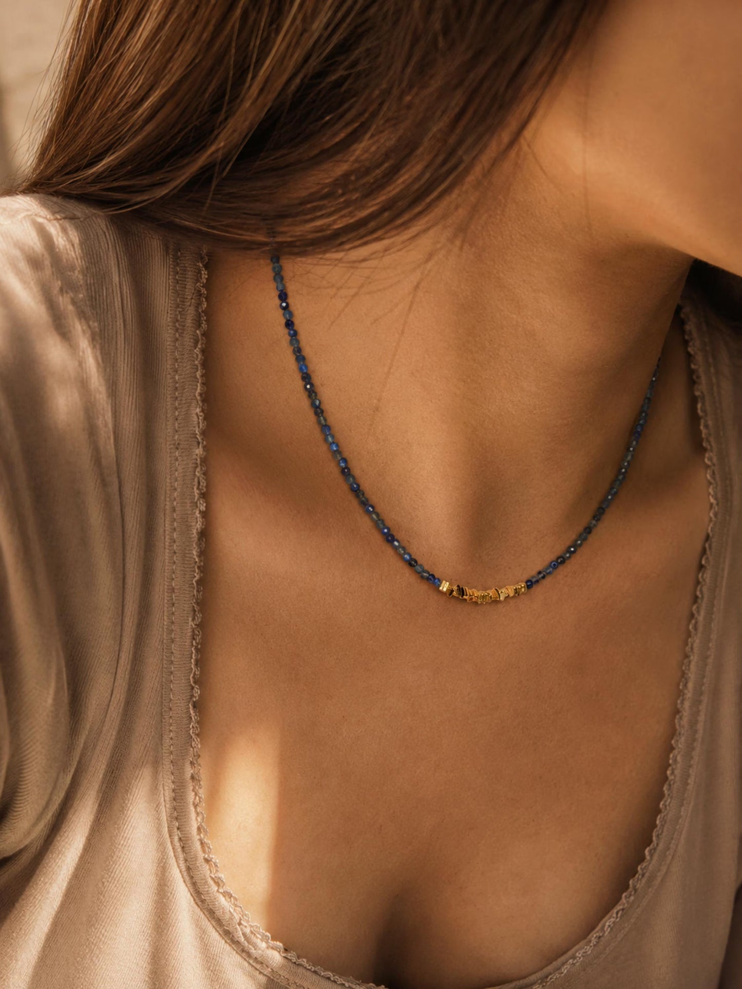 A close-up lifestyle shot of a woman wearing a delicate royal blue beaded necklace with dark blue kyanite beads and a central gold-toned bead accent, paired with a beige satin top.