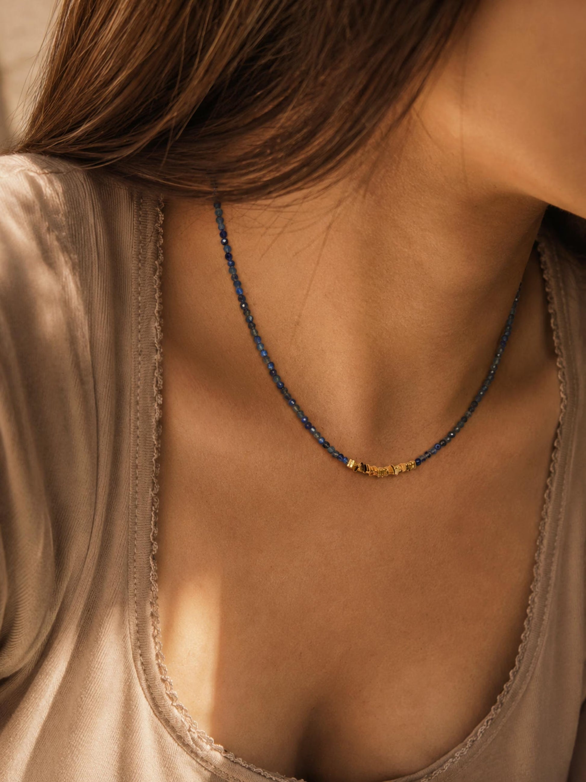 A close-up lifestyle shot of a woman wearing a delicate royal blue beaded necklace with dark blue kyanite beads and a central gold-toned bead accent, paired with a beige satin top.