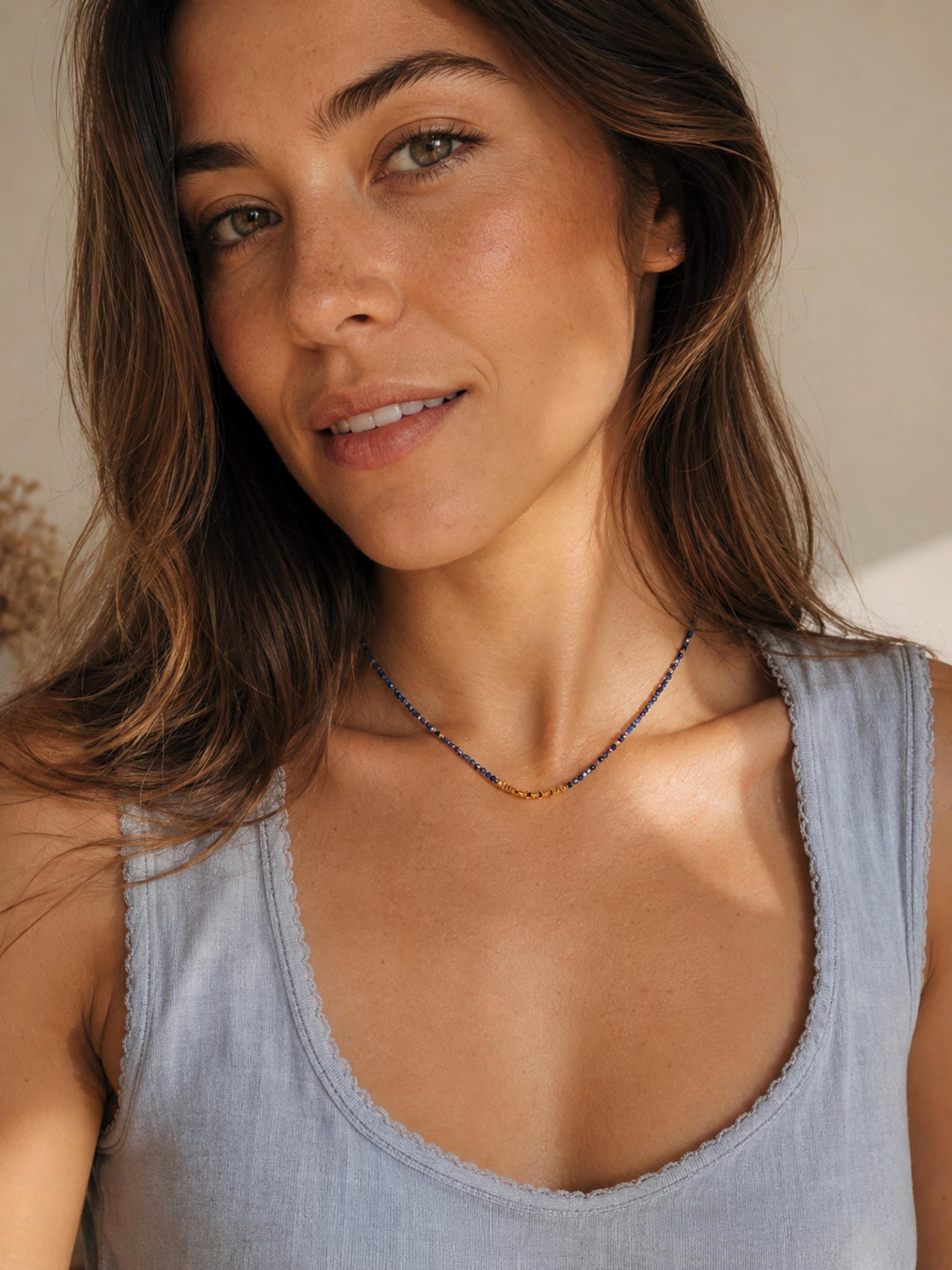 A close-up lifestyle shot of a woman with long brown hair wearing a delicate beaded Kyanite necklace with blue and gold beads, paired with a light blue-gray ribbed tank top.