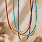 A set of multi-strand beaded necklaces draped in front of a textured beige background with soft shadows and decorative stones. The necklaces feature faceted beads in red, turquoise blue, dark blue, light green, pale pink, and metallic gold.