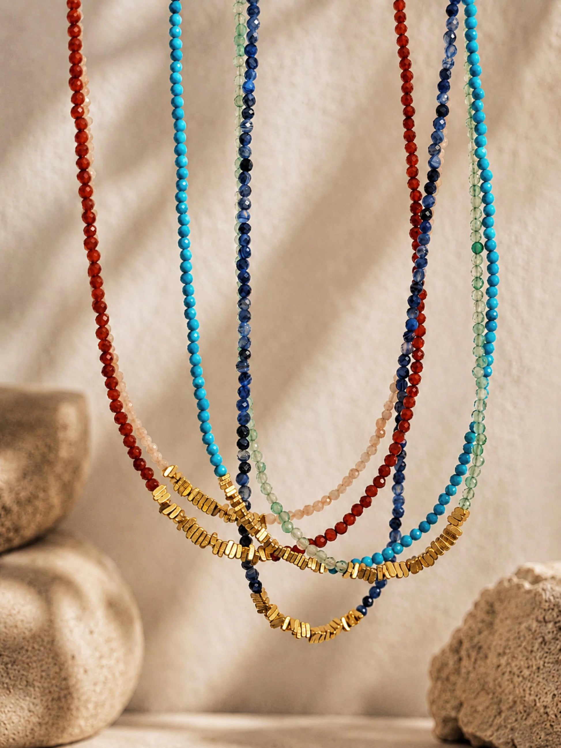 A set of multi-strand beaded necklaces draped in front of a textured beige background with soft shadows and decorative stones. The necklaces feature faceted beads in red, turquoise blue, dark blue, light green, pale pink, and metallic gold.
