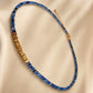 A delicate beaded necklace with alternating strands of faceted blue Kyanite beads and textured gold plated sterling silver beads, displayed on a draped beige satin fabric.
