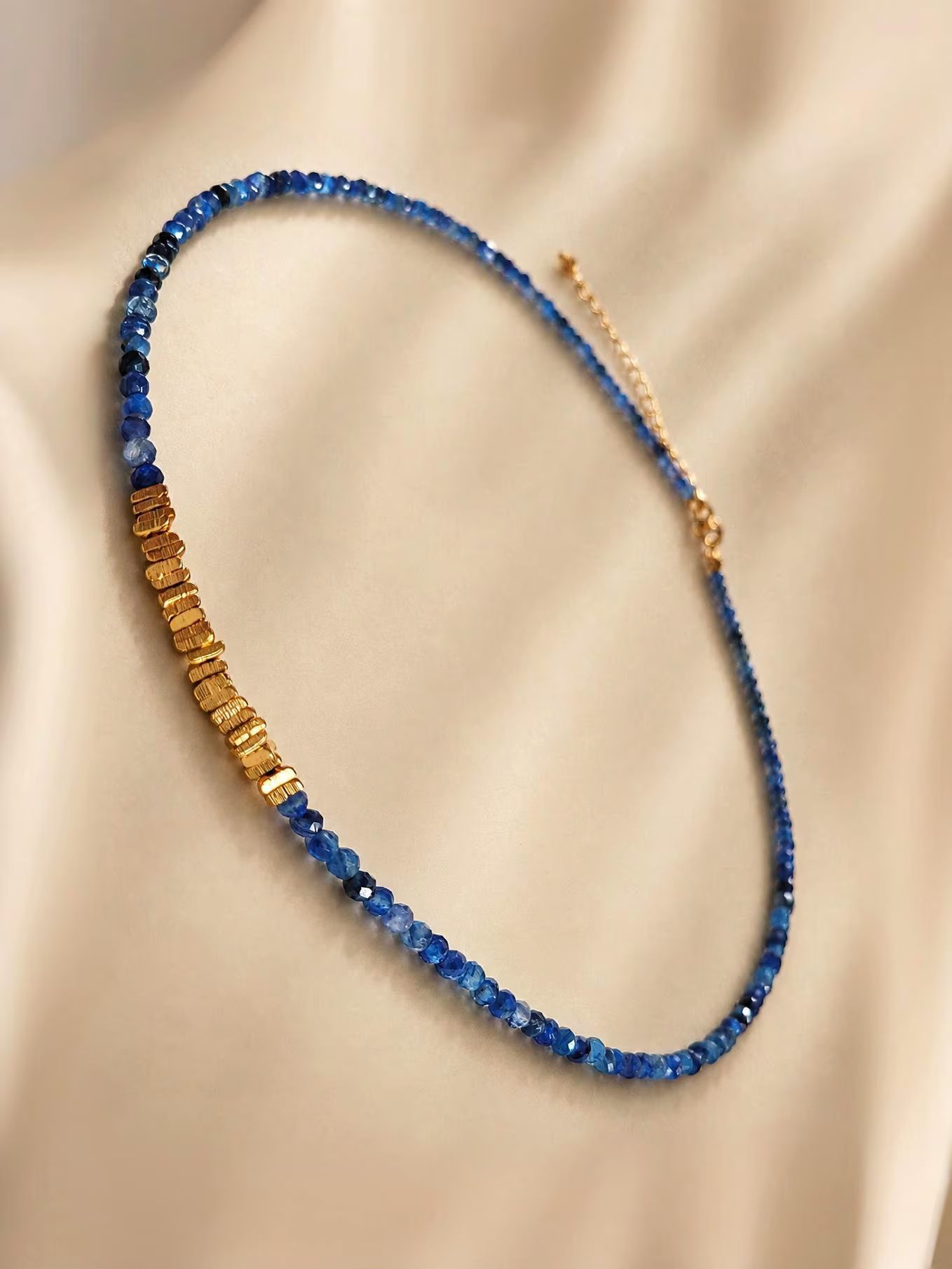 A delicate beaded necklace with alternating strands of faceted blue Kyanite beads and textured gold plated sterling silver beads, displayed on a draped beige satin fabric.