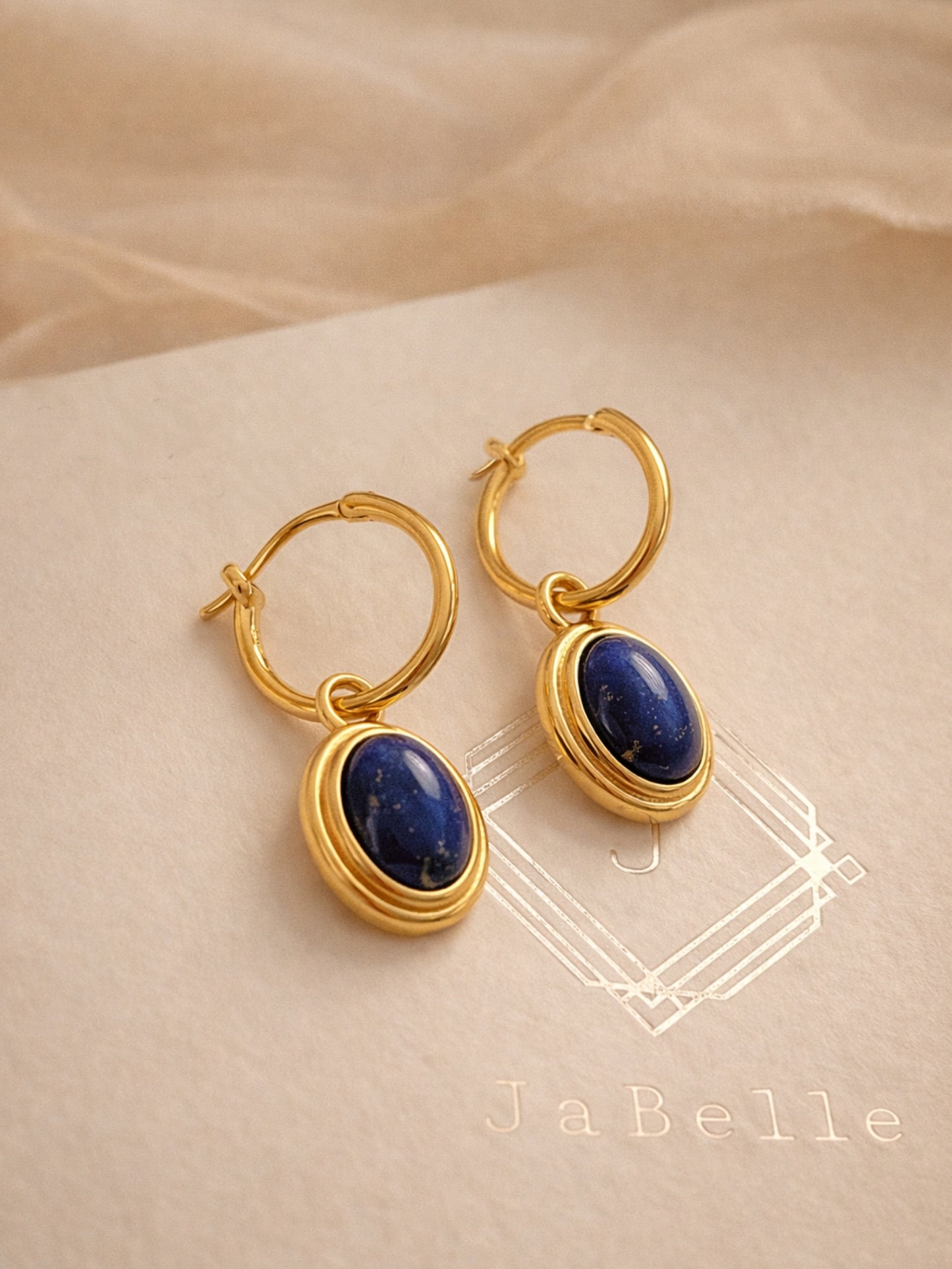 A pair of dainty gold hoop earrings with blue lapiz lazuli gemstone on a beige fabric background.
