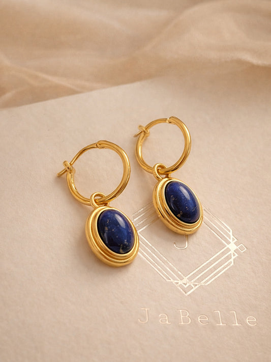 A pair of dainty gold hoop earrings with blue lapiz lazuli gemstone on a beige fabric background.