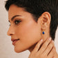 Woman wearing a gold dangle earring with a blue lapis gemstone on a neutral background, elegant and edgy.