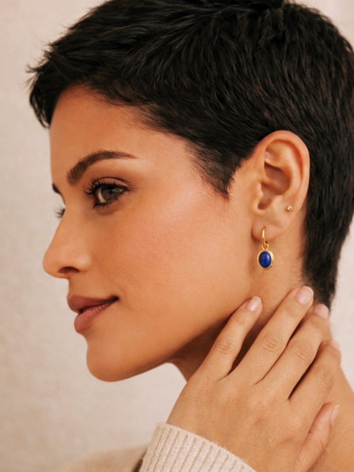 Woman wearing a gold dangle earring with a blue lapis gemstone on a neutral background, elegant and edgy.