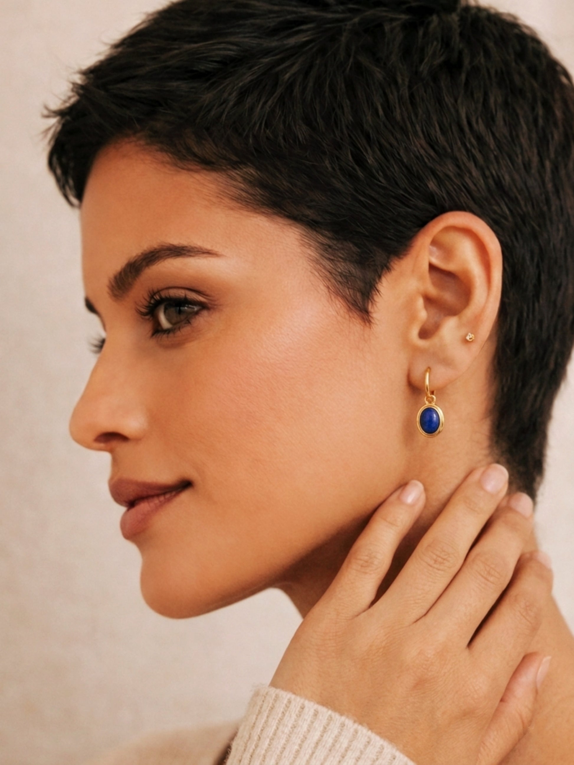 Woman wearing a gold dangle earring with a blue lapis gemstone on a neutral background, elegant and edgy.
