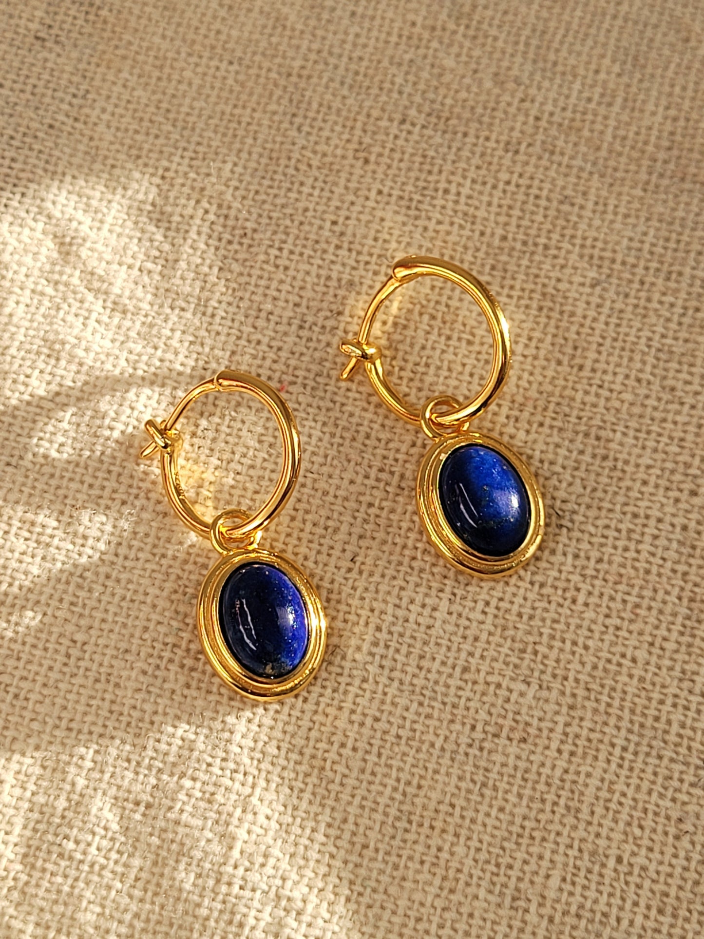 A pair of gold hoop earrings with blue lapis lazuli gemstone, the september birthstone, on a beige fabric.