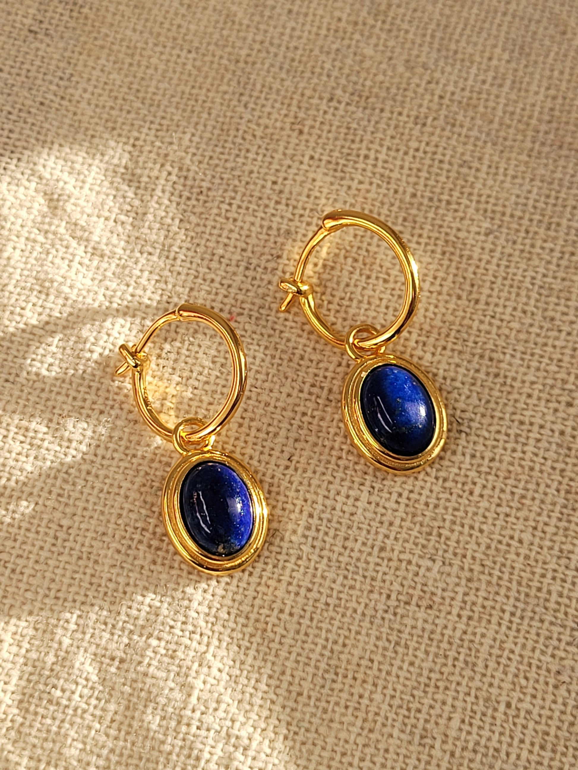 A pair of gold hoop earrings with blue lapis lazuli gemstone, the september birthstone, on a beige fabric.