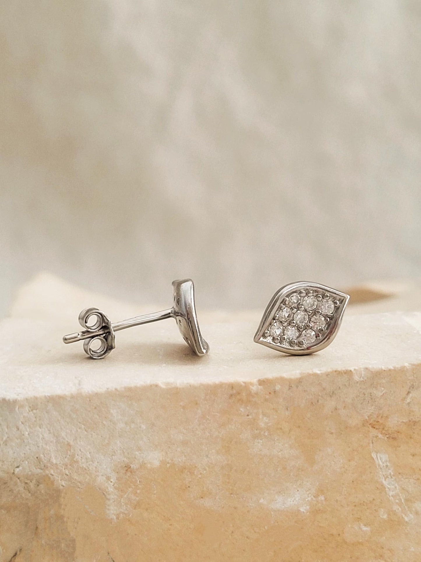 A pair of sculptural leaf cluster earrings displayed on a aesthetic beige stone surgace, showing the design in 925 sterling silver with pave cubic zirconia details.