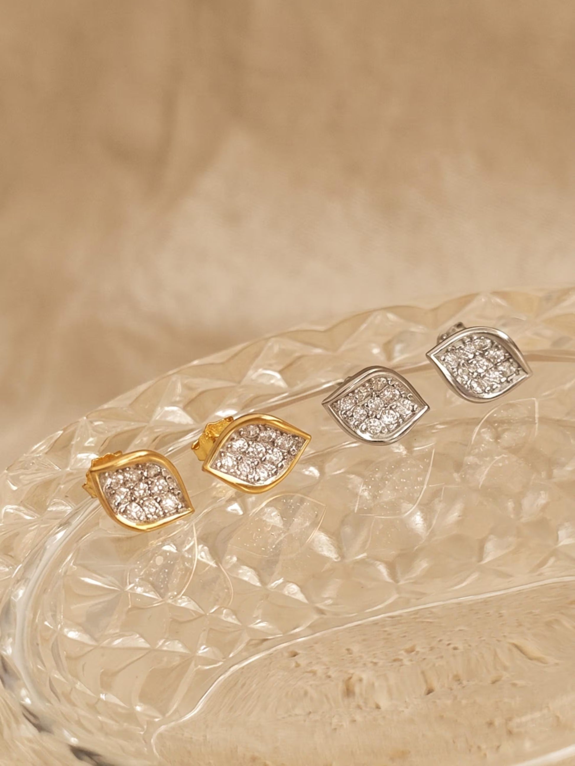 A collection of four sculptural leaf cluster earrings displayed on a glass surface, showing the design in both 18k gold vermeil and 925 sterling silver with pave cubic zirconia details.