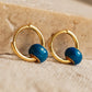 Close up of a pair of mini blue enamel bead huggie earrings crafted in gold plated sterling silver with a small enamel bead, displayed on an aesthetic beige color surface, showcasing its dainty and minimalist details.