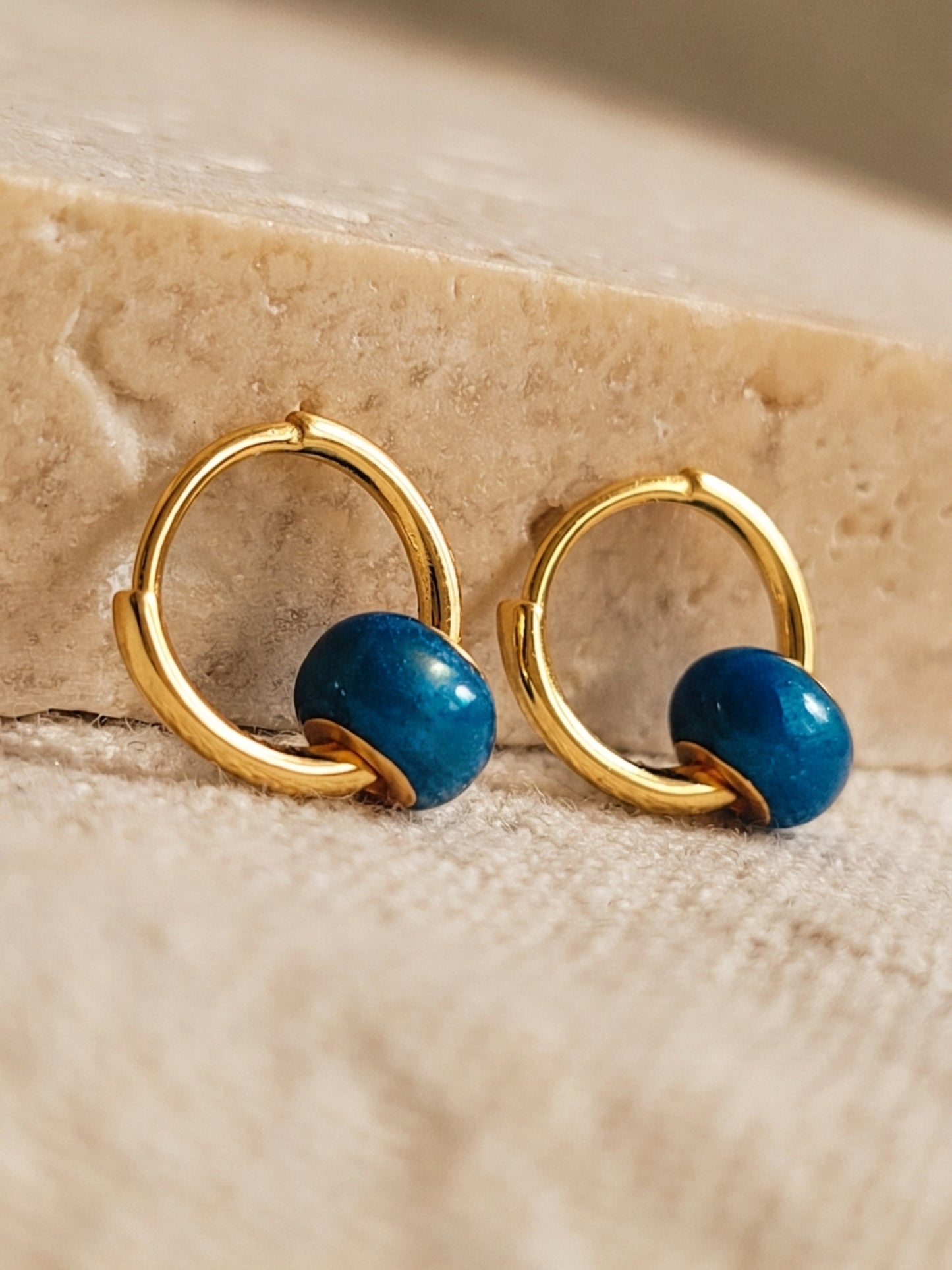 Close up of a pair of mini blue enamel bead huggie earrings crafted in gold plated sterling silver with a small enamel bead, displayed on an aesthetic beige color surface, showcasing its dainty and minimalist details.