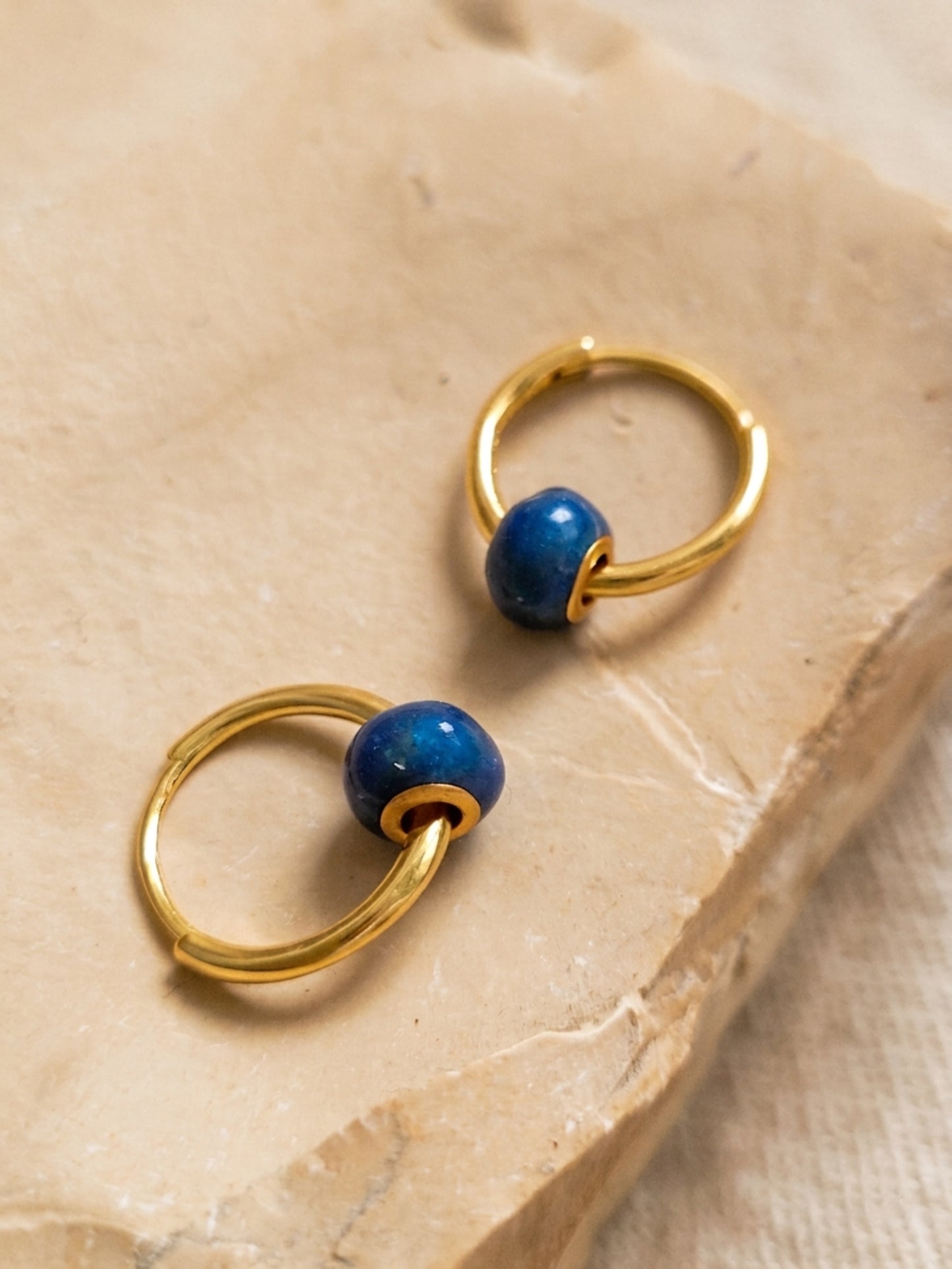 A pair of mini blue enamel bead huggie earrings displayed on an aesthetic beige color stone surface, showcasing its dainty and minimalist details.
