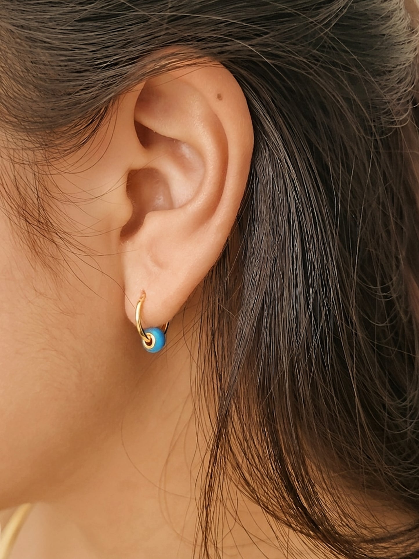 Close-up of an ear wearing a gold hoop earring with a blue enamel bead, crafted in London UK