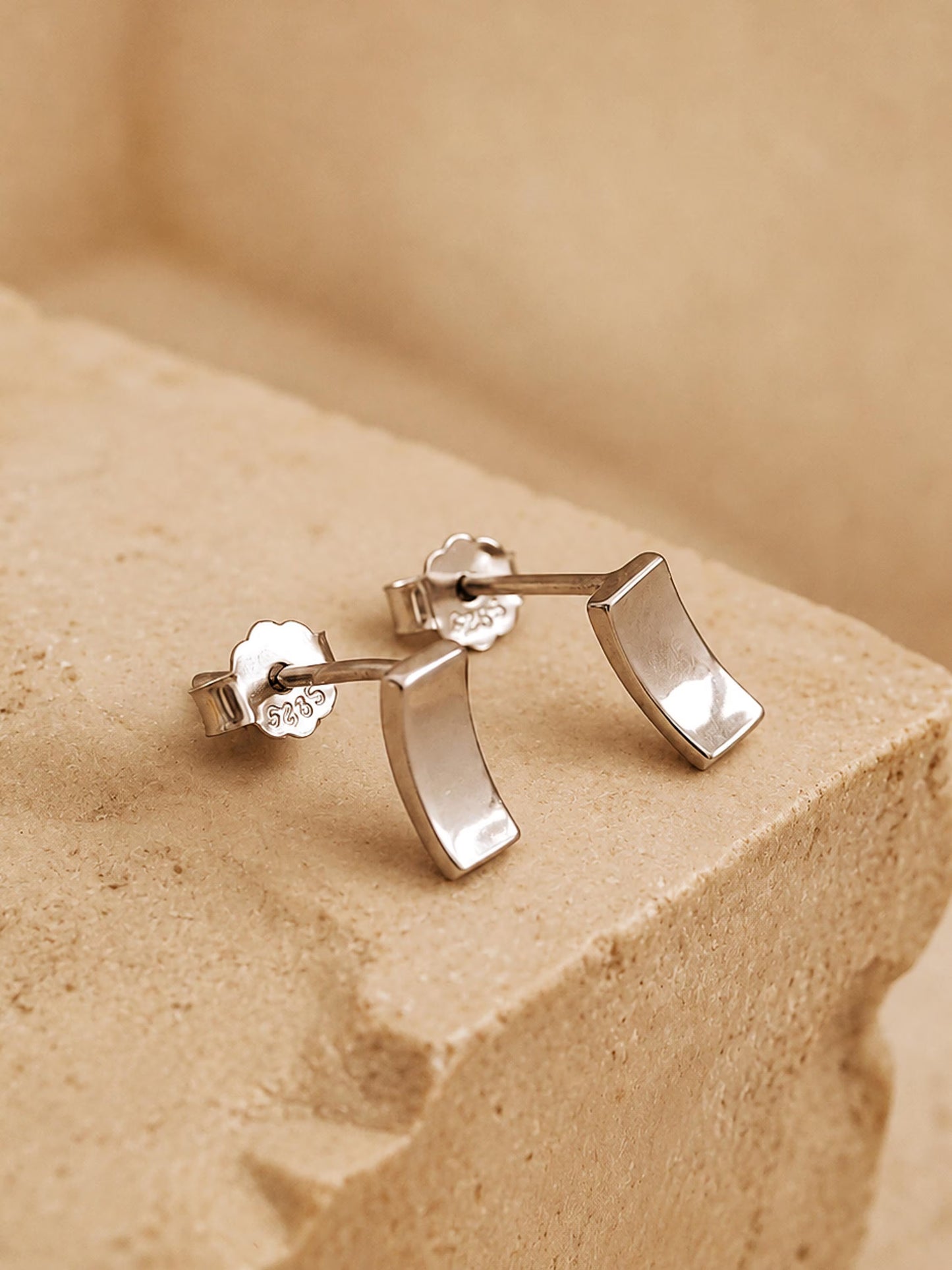 A pair of tiny silver stud earrings in curved bar shape on a beige stone surface, simple and minimal style.