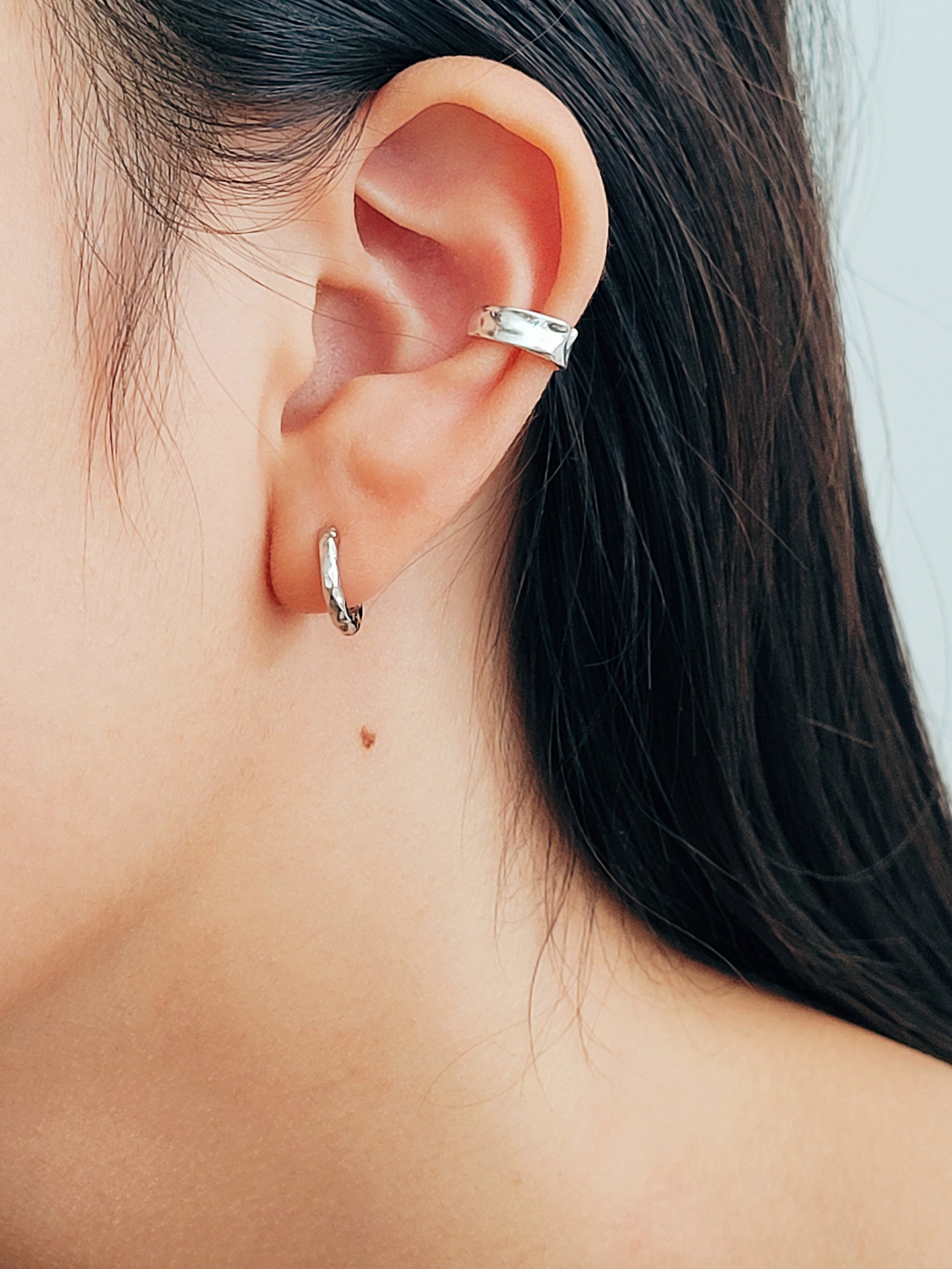 Close-up of an ear wearing a textured silver ear cuff earring paired with a simple hoop earrings with a neutral background