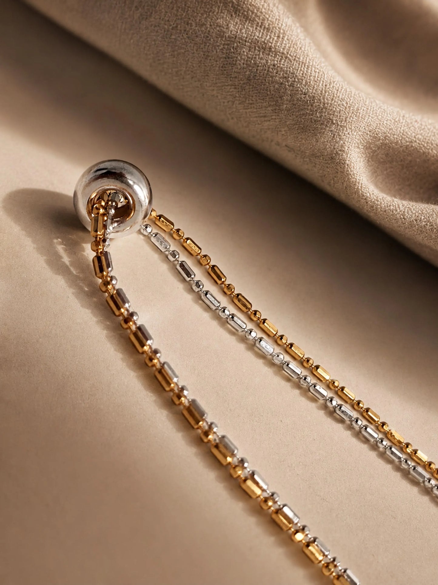 Close-up shot of a two-tone gold and silver beaded double chain necklace with a small silver ring charm resting on soft textured linen fabric.
