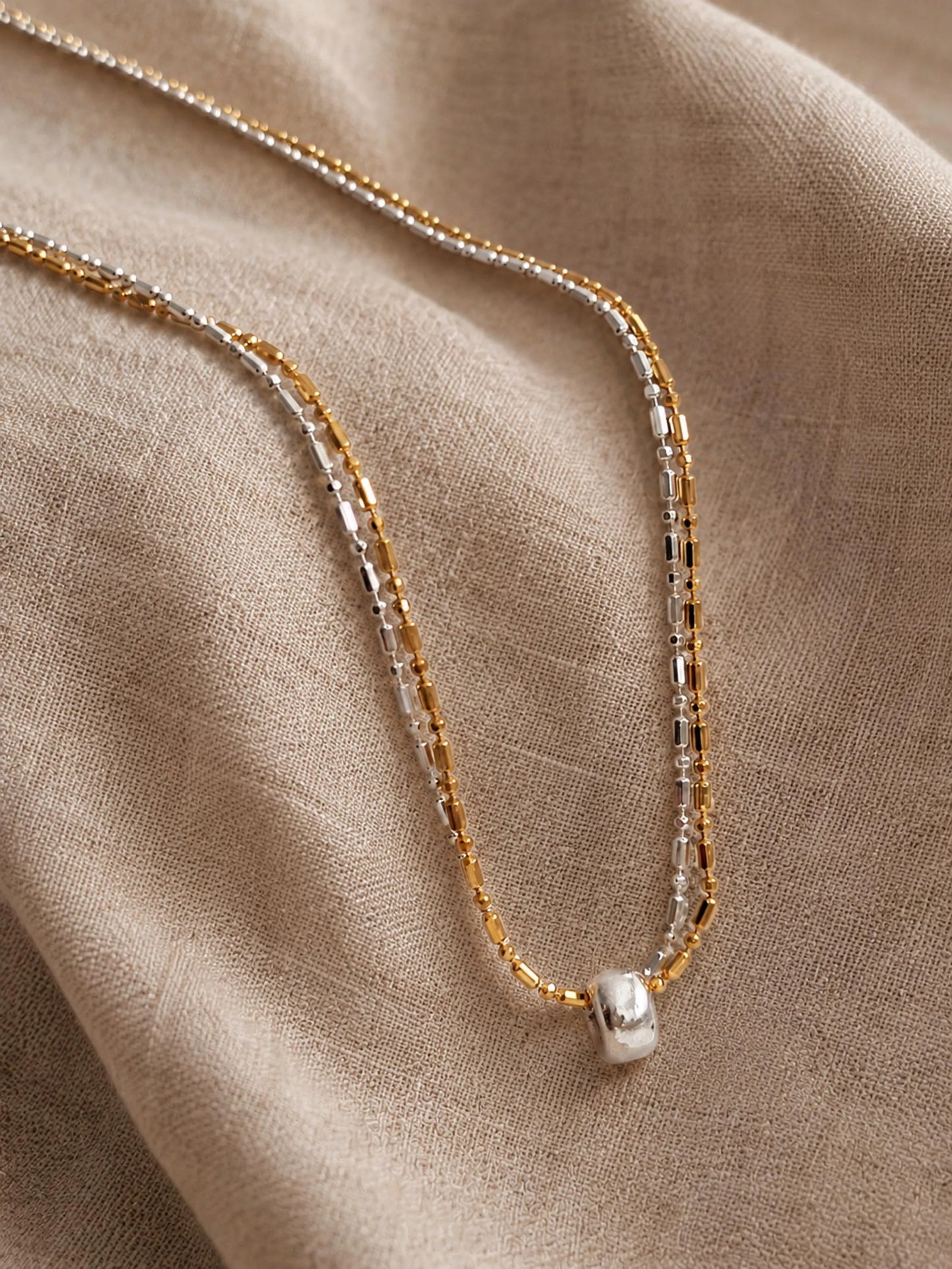 A delicate double-strand necklace with one gold and one silver chain, featuring a small silver ring charm, displayed on a textured beige linen fabric.