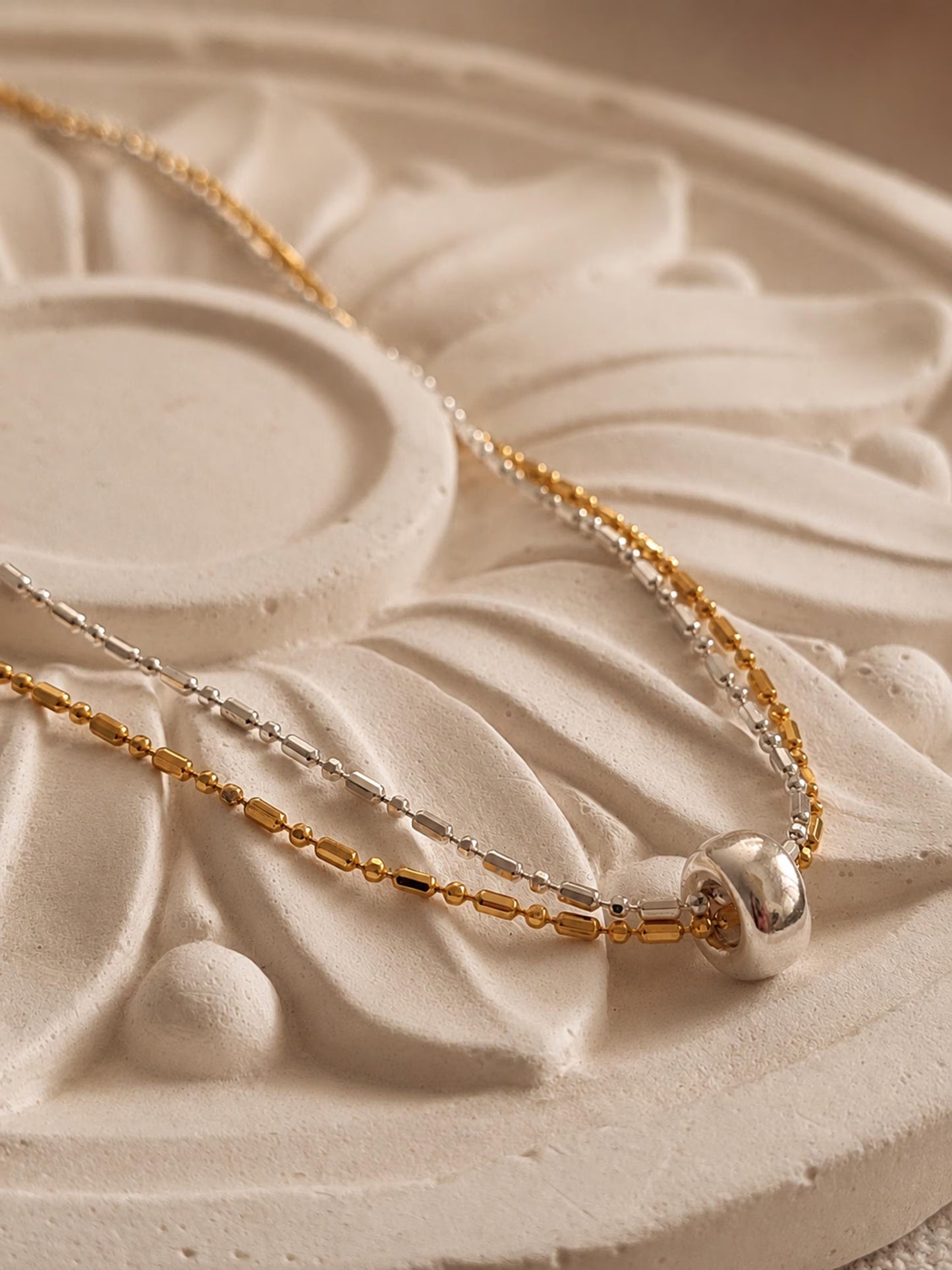 A close-up of a delicate two-tone necklace with gold and silver chains, featuring a polished silver bead pendant, resting on a textured beige stone surface.