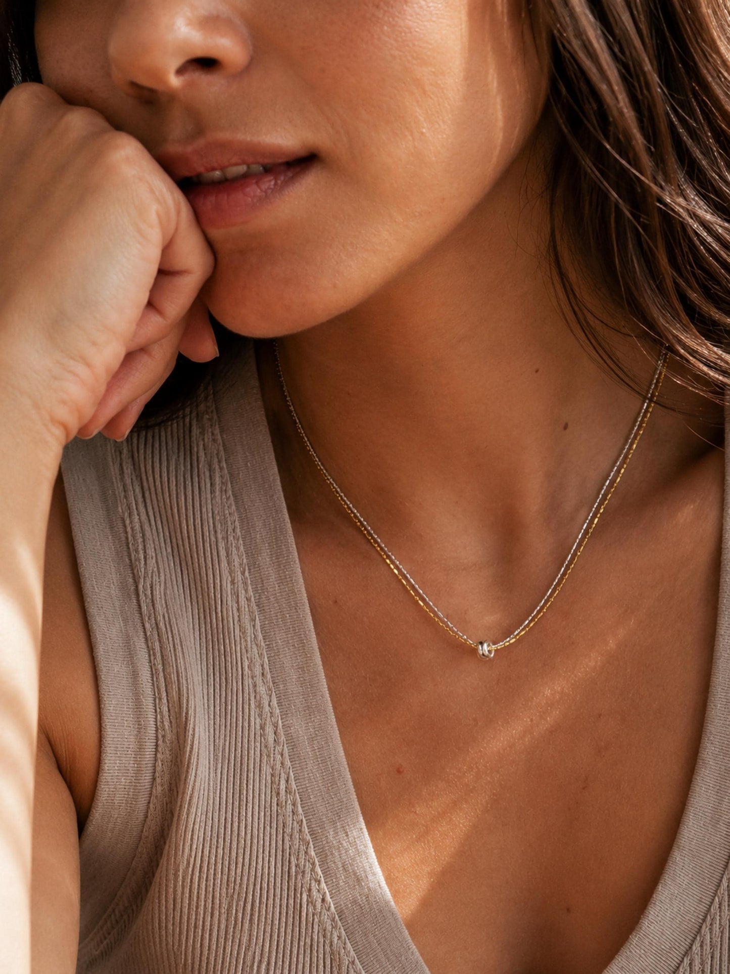 A close-up lifestyle shot of a woman wearing a delicate gold chain necklace with a small round pendant, paired with a beige ribbed top.