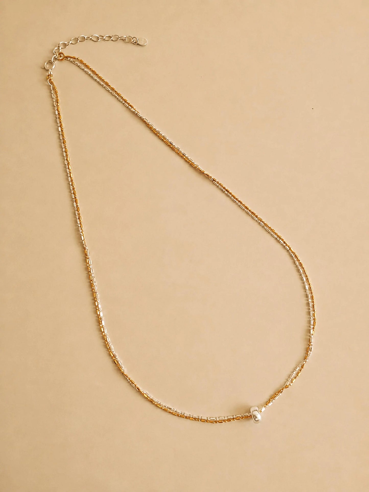 A delicate gold chain necklace with a small silver round pendant, displayed on a solid beige background.