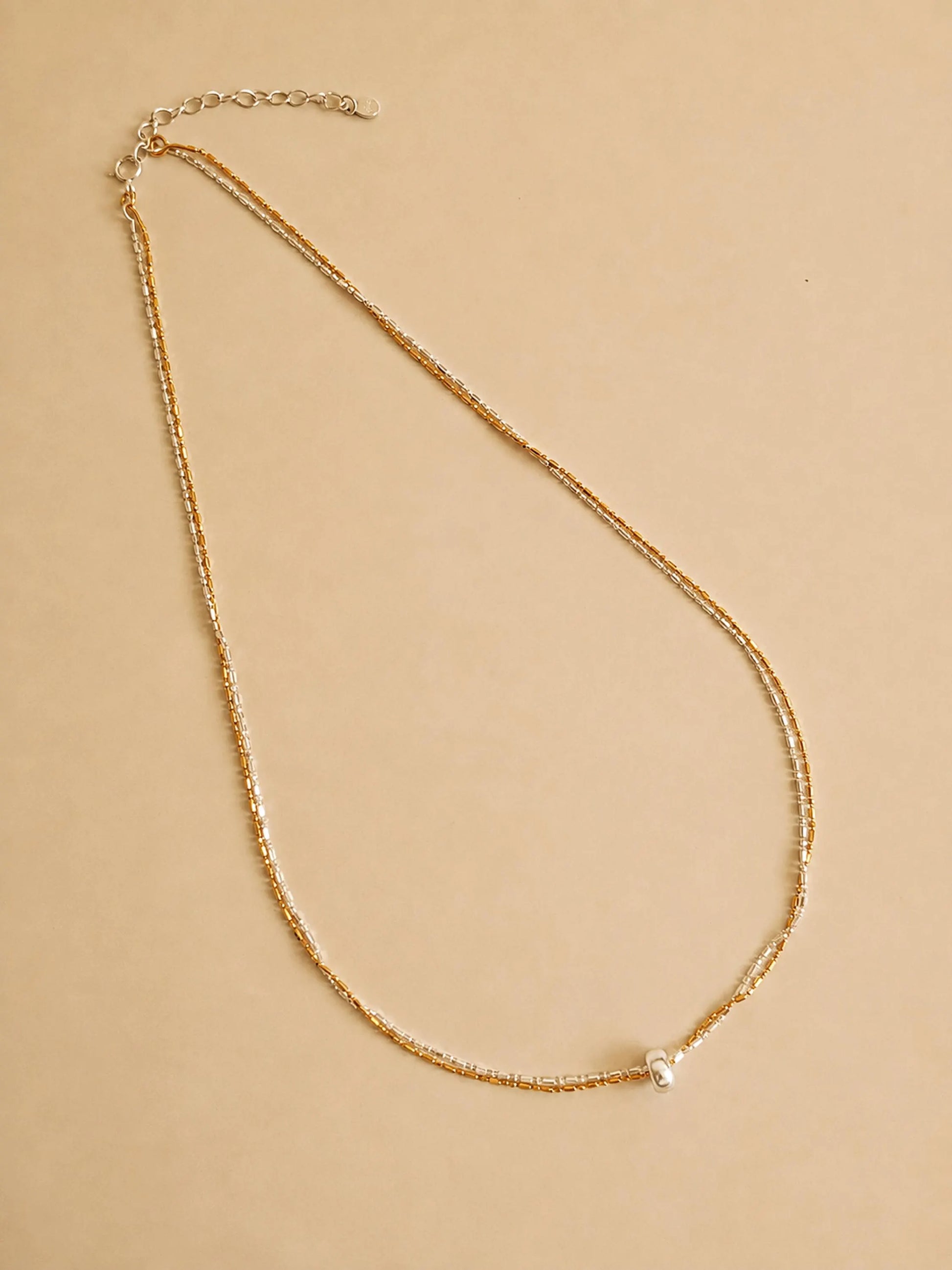 A delicate gold chain necklace with a small silver round pendant, displayed on a solid beige background.