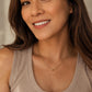 A woman with brown hair wearing a delicate gold chain necklace with a small bead pendant, paired with a beige ribbed tank top.