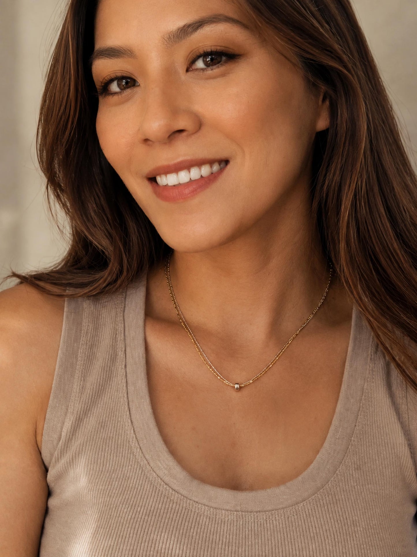 A woman with brown hair wearing a delicate gold chain necklace with a small bead pendant, paired with a beige ribbed tank top.