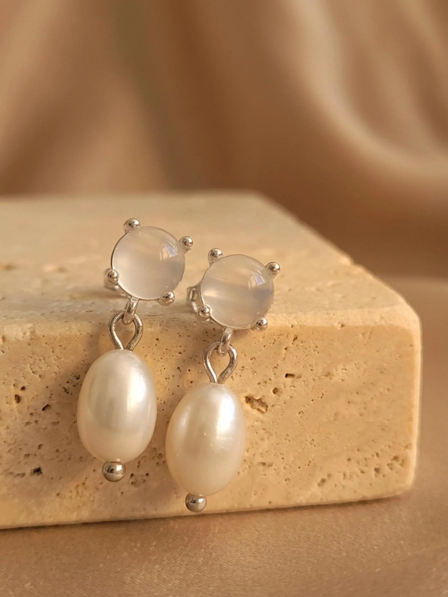 A pair of dainty silver stud earrings with moonstone and freshwater pearl drop crafted in 925 sterling silver displayed on an aesthetic beige color stone, minimal wedding earrings.