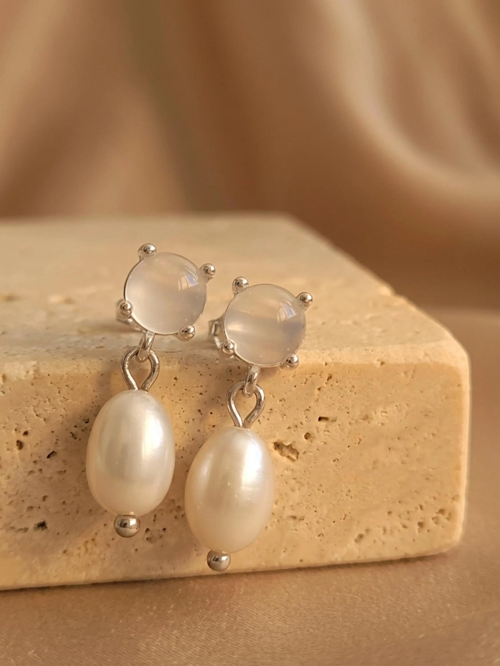 A pair of dainty silver stud earrings with moonstone and freshwater pearl drop crafted in 925 sterling silver displayed on an aesthetic beige color stone, minimal wedding earrings.
