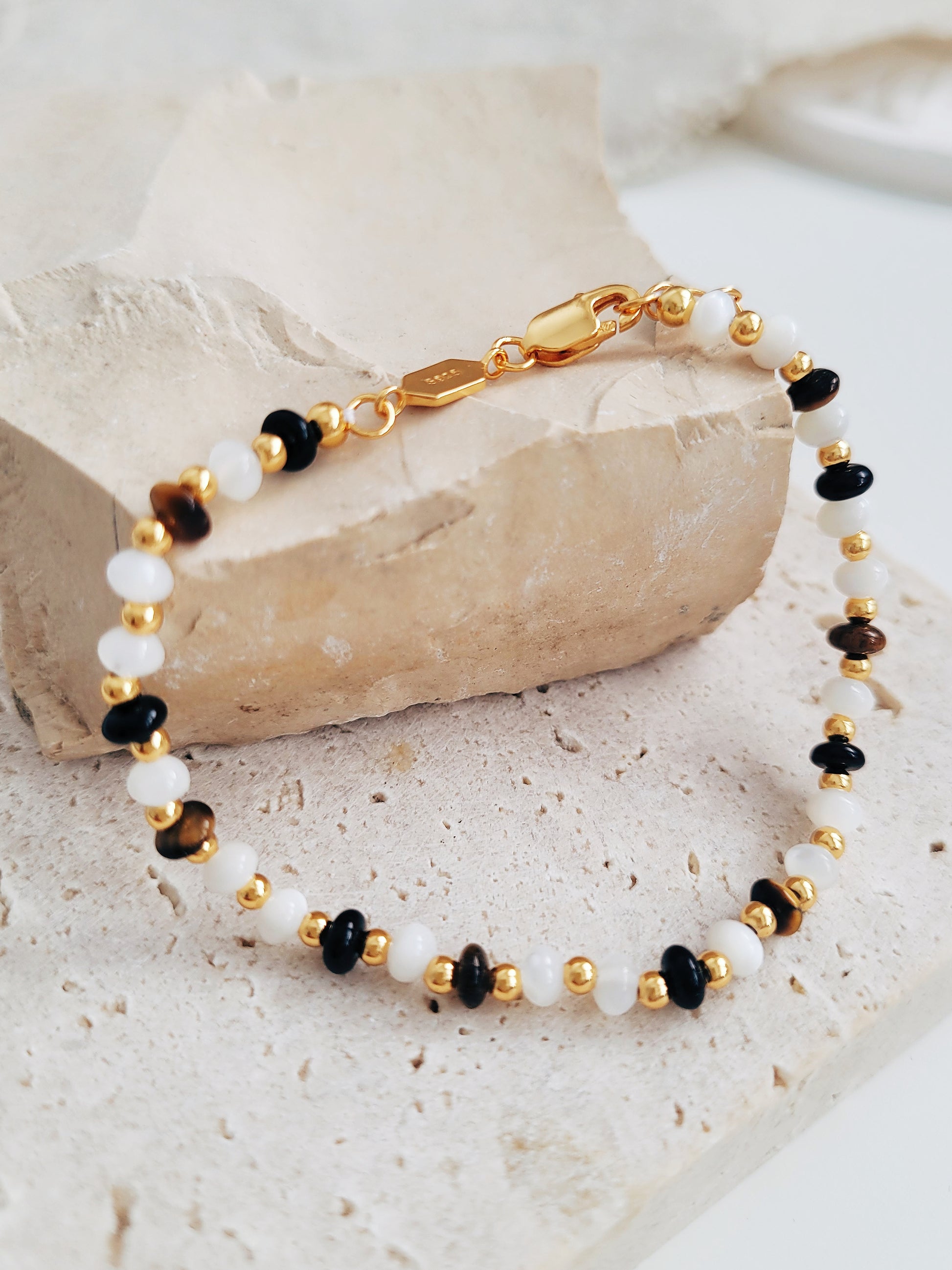 A natural stone bracelet featuring tiger's eye, mother of pearl, and black onyx beads with 22k gold vermeil accents lied on a stone