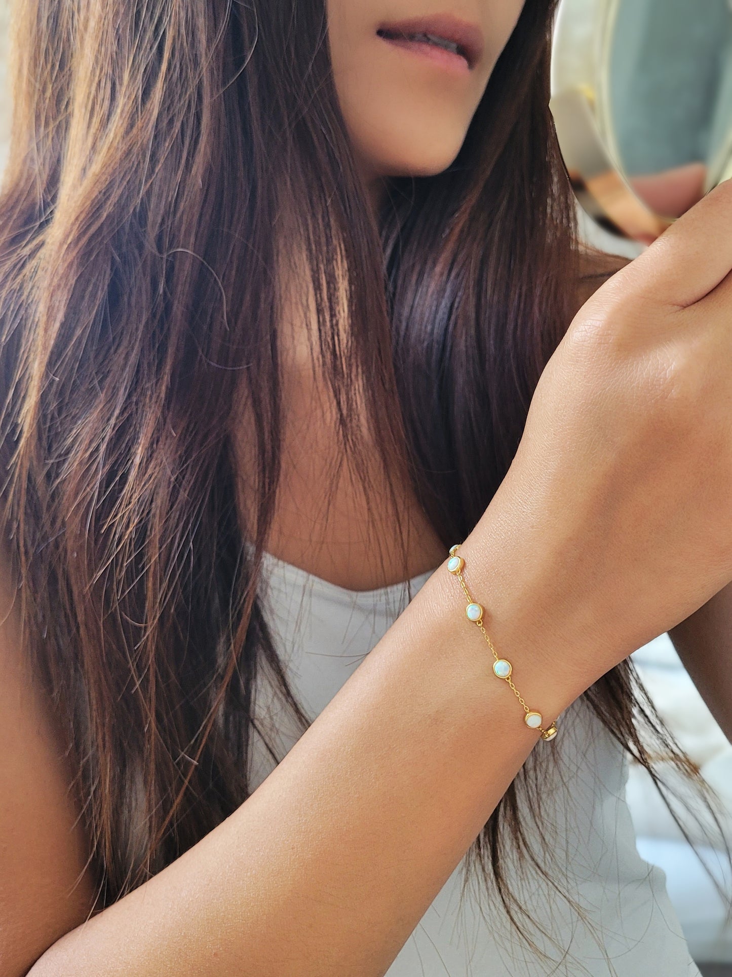 A woman elegantly holds a mirror, showcasing her stunning gold-plated opal bracelet with white opal stones.