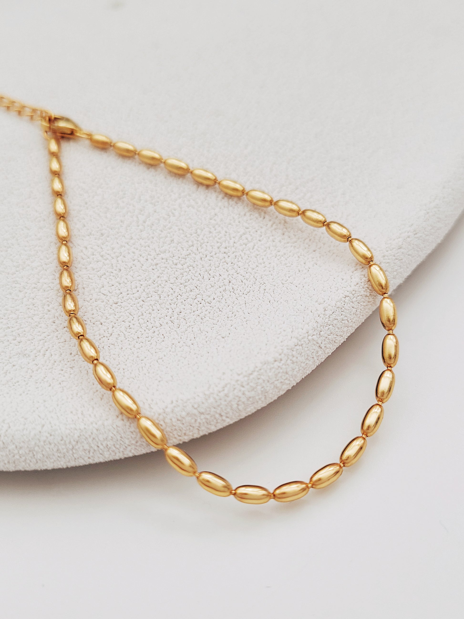 A simple and delicate gold-plated beaded bracelet is placed on a neutral background featuring a series of shimmering oval shaped beads, highlighting its lustrous finish and intricate bead arrangement.