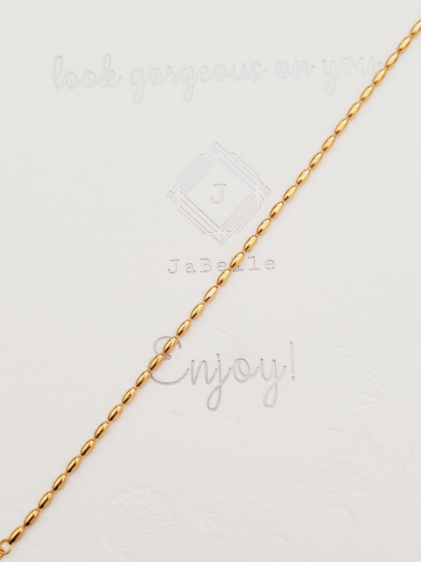 A simple and delicate gold-plated beaded bracelet is placed on a neutral background featuring a series of shimmering oval shaped beads, highlighting its lustrous finish and intricate bead arrangement.