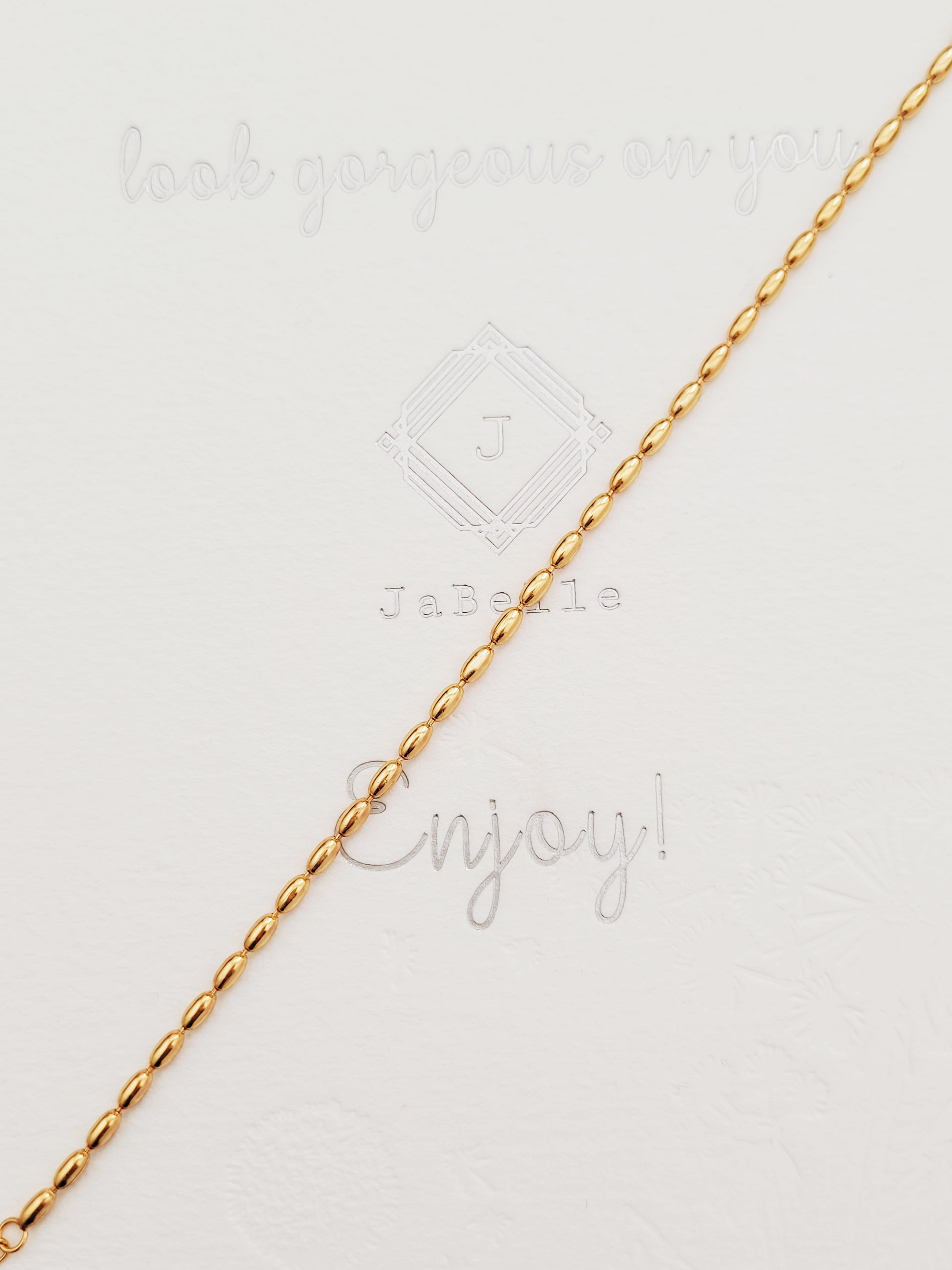 A simple and delicate gold-plated beaded bracelet is placed on a neutral background featuring a series of shimmering oval shaped beads, highlighting its lustrous finish and intricate bead arrangement.