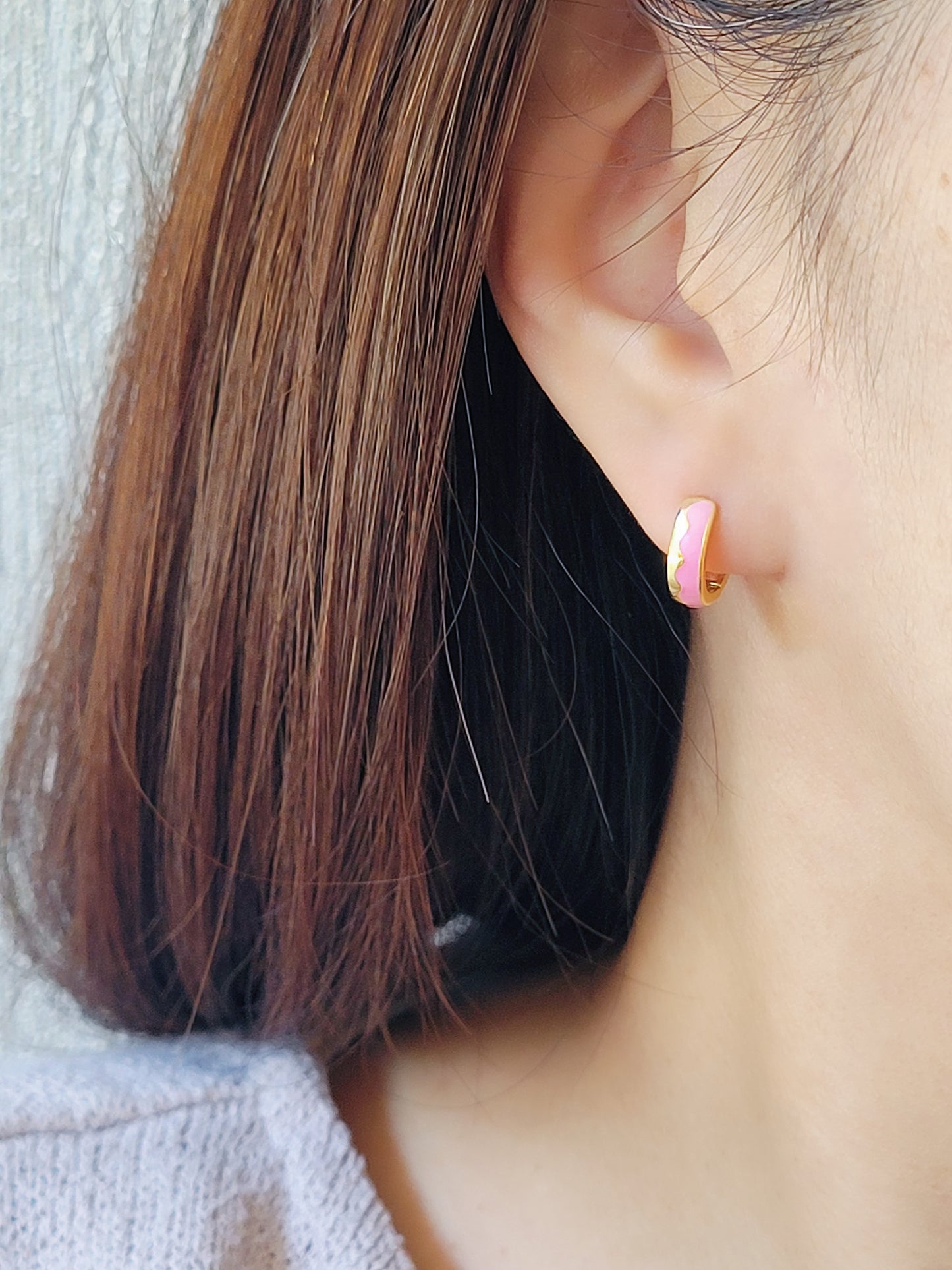 Close-up of a woman’s ear wearing pink and gold enamel huggie earrings, showcasing their smooth finish and playful wavy design.